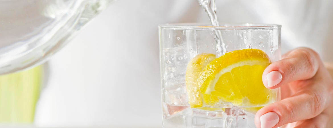 A person pours water from a pitcher into a glass with a lemon slice in it