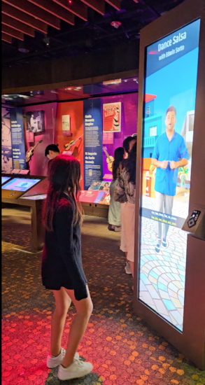 A girl follows instructions on how to salsa dance from a salsa teacher portrayed on a large video screen.
